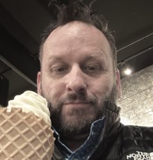 A white man with brown hair, a beard, and mustache holding an ice cream cone looks at the camera.