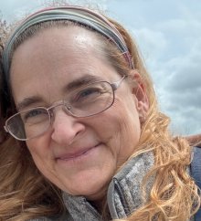 A white woman with glasses and blonde hair held back by a headband smiles at the camera.