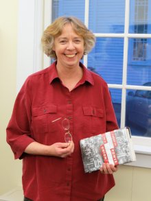 A white woman in a red shirt smiles at the camera. She has a book in one hand and her glasses in another.